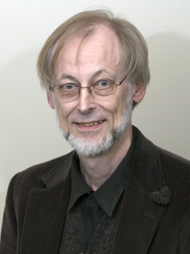 An older man with light skin and hair poses before a neutral backdrop smiling and wearing a brown corduroy sport coat and dark tie and shirt