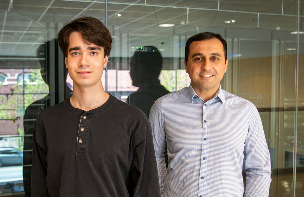 Two men with dark hair standing in front of a glass wall housing computer servers